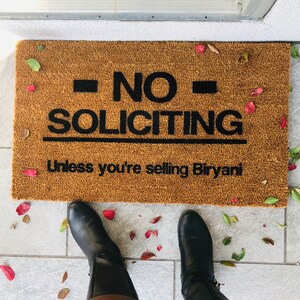 Puede incluir: Una alfombra de bienvenida marr&oacute;n con el texto "NO SOLICITING Unless you're selling Biryani" en letras negras. La alfombra est&aacute; sobre un suelo de baldosas con hojas ca&iacute;das.