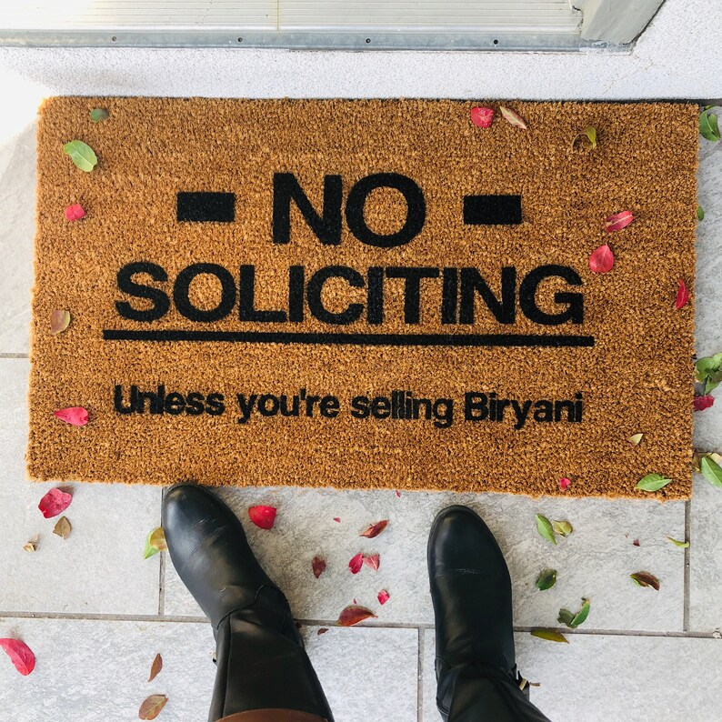Puede incluir: Una alfombra de bienvenida marr&oacute;n con el texto "NO SOLICITING Unless you're selling Biryani" en letras negras. La alfombra est&aacute; sobre un suelo de baldosas con hojas ca&iacute;das.