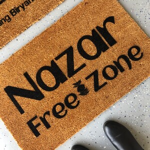 Puede incluir: Una alfombra de coco marr&oacute;n con el texto "Nazar Free Zone" en letras negras.