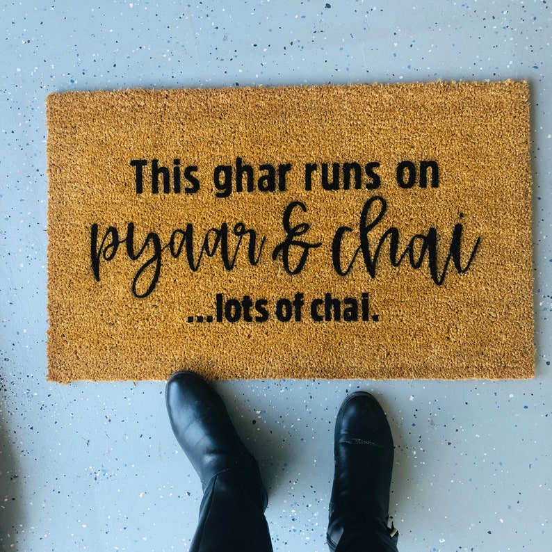 Puede incluir: Una alfombra de coco marr&oacute;n con el texto "This ghar runs on pyaar & chai...lots of chai." en letras negras.