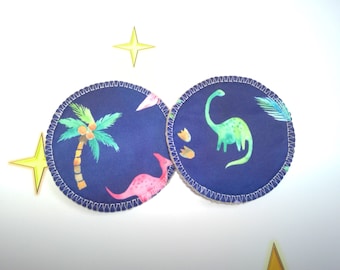 May include: Two round, blue fabric coasters with white stitching. One coaster features a pink dinosaur and a palm tree with coconuts. The other coaster features a green dinosaur and footprints.