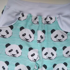 Op de afbeelding: Tealblauwe stof met een witte tailleband en witte zakken. De stof heeft een herhalend patroon van zwart-witte panda gezichten.