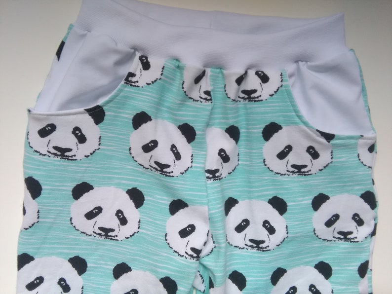 Op de afbeelding: Tealblauwe stof met een witte tailleband en witte zakken. De stof heeft een herhalend patroon van zwart-witte panda gezichten.