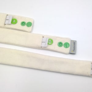 Op de afbeelding: Drie crèmekleurige stoffen banden met groene drukkers en elastische lussen. De banden hebben een zacht, getextureerd uiterlijk. Een klein label met tekst is zichtbaar op een band.
