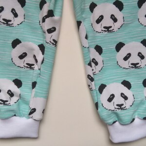 Op de afbeelding: Een paar tealblauwe sweatpants met een witte en zwarte pandabeerprint. De sweatpants hebben witte elastische boorden aan de onderkant.