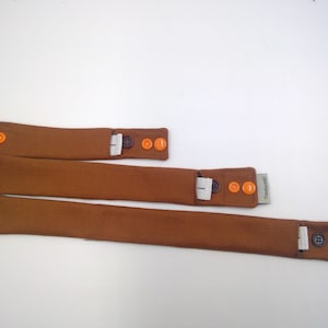 Könnte beinhalten: Drei braune Stoffarmbänder mit orangefarbenen Knöpfen und einer weißen Tasche. Die Armbänder sind in verschiedenen Größen.