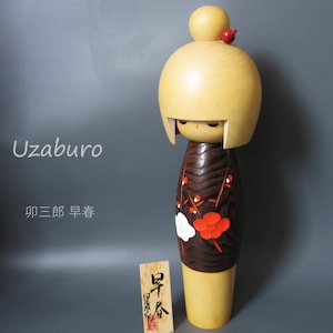 May include: A wooden Kokeshi doll with a light-colored rounded head and a dark brown body featuring painted red and white plum blossoms. The doll has a small red accent on top of its head. Text on the left reads Uzaburo and 卯三郎 早春.