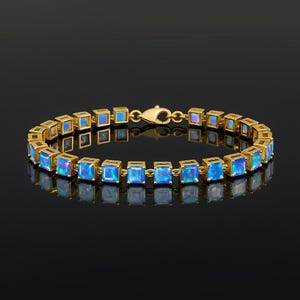 Op de afbeelding: Een goudkleurige armband met vierkante, iriserende blauwe edelstenen. De armband bestaat uit afzonderlijke vierkante schakels, elk met een edelsteen. De sluiting is ook goudkleurig. De armband wordt tegen een donkere achtergrond getoond.