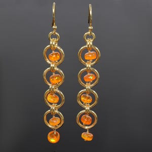 May include: A pair of long, dangling earrings. Each earring has gold-coloured rings connected by smaller gold links. Orange-coloured, oval-shaped beads are suspended within the rings and at the bottom of each earring. The earrings have a hook closure.