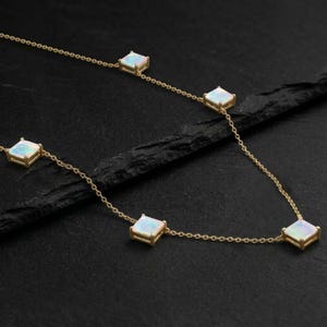 May include: A delicate gold necklace featuring five square-cut opal stones. The opals are set in gold bezels and connected by a fine gold chain. The necklace is displayed against a dark background, highlighting the jewelry's sparkle.