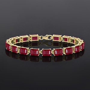 May include: A gold bracelet featuring rectangular, deep red gemstones. The bracelet is composed of individual links, each holding a ruby-colored stone. The clasp is also gold, and the bracelet is displayed against a dark background.