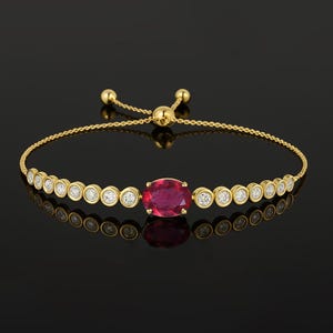 May include: A gold bracelet with a large oval ruby centre stone, surrounded by smaller round diamonds in bezel settings. The bracelet has a fine chain and a sliding bead closure.