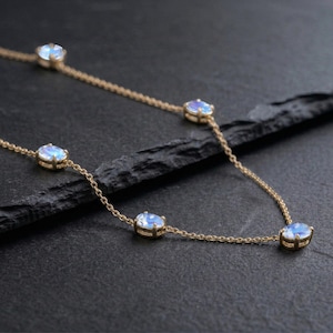 May include: A delicate gold necklace featuring oval-shaped, iridescent gemstones. The gemstones are evenly spaced along a fine gold chain. The necklace is displayed on a dark, textured surface, highlighting the jewelry's subtle sparkle.