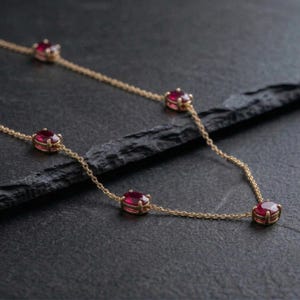 May include: A delicate gold necklace featuring oval-shaped red gemstones. The gemstones are evenly spaced along the fine chain. The necklace is displayed on a dark, textured surface, highlighting the jewelry's elegance.