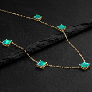 May include: A delicate gold necklace featuring square-cut, iridescent green gemstones. The gemstones are set in gold bezels and linked by a fine gold chain. The necklace is displayed on a dark, textured surface.
