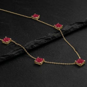 May include: A delicate gold necklace featuring square-cut, vibrant red gemstones. The gemstones are evenly spaced along the fine chain, creating a sophisticated and elegant design. The necklace is displayed against a dark, textured background.
