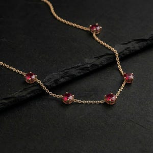 May include: A gold necklace with five red gemstones. The necklace is set against a dark backdrop, accentuating the contrast between the gold chain and the red stones. The gemstones are evenly spaced along the chain, creating a balanced design.