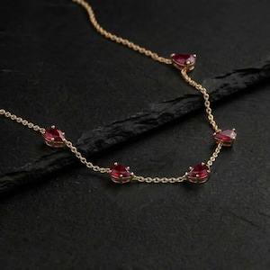 May include: A delicate gold necklace featuring teardrop-shaped red gemstones. The necklace has a fine chain and is set against a dark background, highlighting the vibrant red of the stones. A piece of jewelry for everyday wear or special occasions.