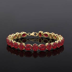 May include: A gold bracelet with oval-cut red gemstones. The bracelet has a classic design with each gemstone set in a gold bezel. The clasp is also gold. The bracelet is displayed on a reflective black surface.
