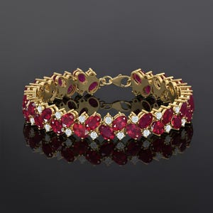May include: A gold bracelet with oval-cut red gemstones and small white gemstones. The bracelet has a clasp closure and is displayed on a reflective surface. The gemstones are arranged in a repeating pattern.