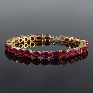 Könnte beinhalten: Ein goldenes Armband mit einer Reihe leuchtend roter Edelsteine in verschiedenen Formen, darunter ovale, birnenförmige und rechteckige Schliffe. Das Armband ist vor einem dunklen Hintergrund platziert, was das Funkeln der Edelsteine hervorhebt.