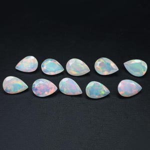 May include: Ten pear-shaped white opals with iridescent rainbow colors. The opals are arranged in a row on a black surface.