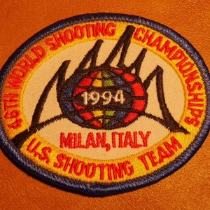 May include: A white oval patch with a blue border and yellow stitching. The patch features a black silhouette of two hands holding a globe with the year 1994 and the text "45th World Shooting Championships U.S. Shooting Team Milan, Italy".