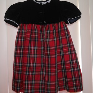 May include: A red and black plaid dress with a black velvet bodice and white lace trim. The dress has a button closure in the back.