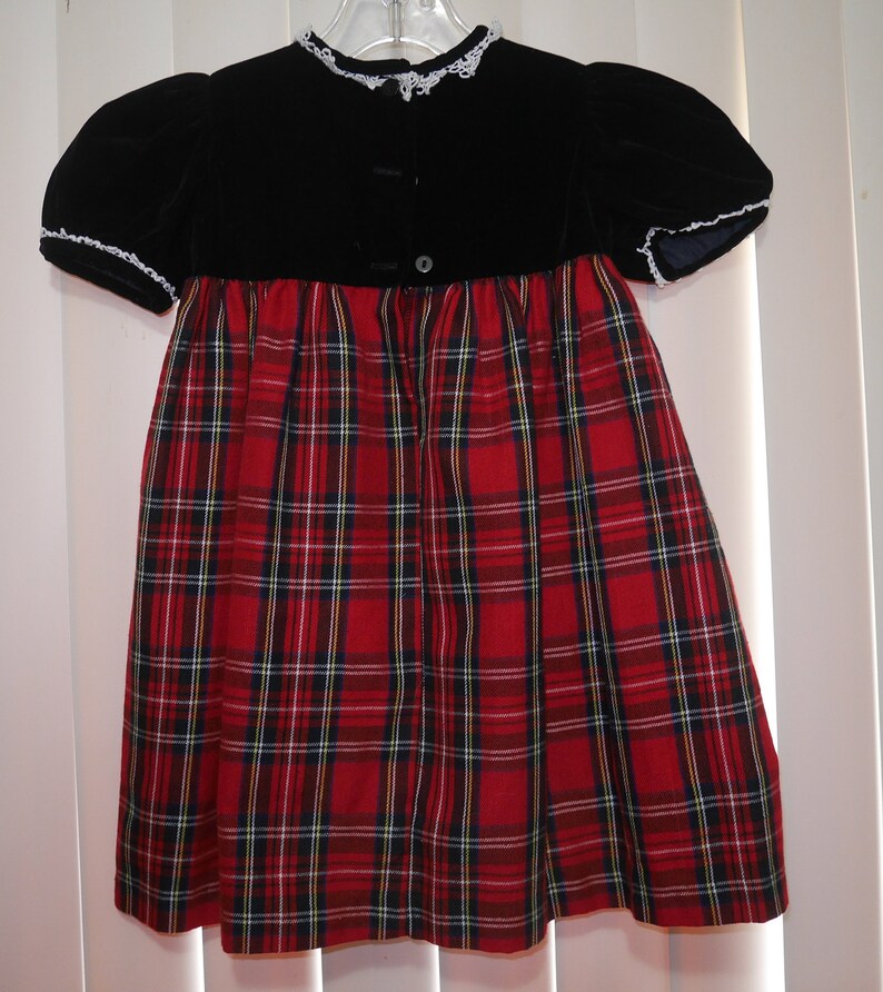 May include: A red and black plaid dress with a black velvet bodice and white lace trim. The dress has a button closure in the back.