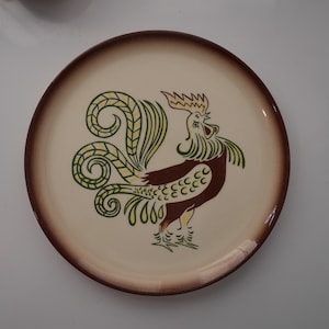 May include: A cream-colored ceramic plate with a brown rim features a stylized green and brown rooster design.