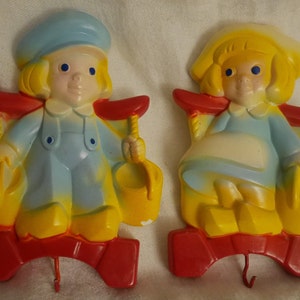 May include: A pair of vintage plastic wall hooks shaped like children. The boy is wearing a blue hat and overalls and holding a yellow bucket. The girl is wearing a yellow dress and holding a yellow bucket. Both are sitting on red rocking chairs.