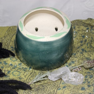 May include: A teal green ceramic planter with a white rim and green leaf design. The planter has two drainage holes.