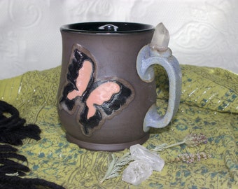 May include: A brown ceramic mug with a butterfly design and a crystal handle. The mug is sitting on a green and white patterned fabric.