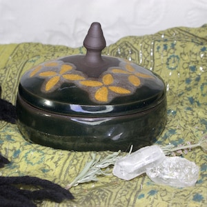 May include: A dark green ceramic box with a lid and a knob on top. The lid is decorated with yellow flower designs. The box is sitting on a green and white patterned fabric.