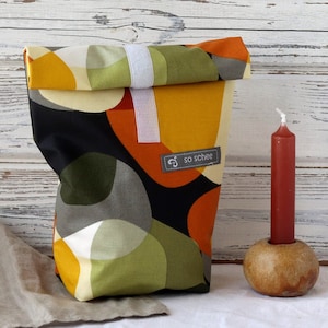 Könnte beinhalten: Eine schwarze und weiße Stoff-Lunch-Tasche mit einem geometrischen Muster in Orange-, Gelb- und Grüntönen. Die Tasche hat ein weißes Etikett mit dem Text "so schee" darauf.