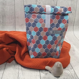 Könnte beinhalten: Eine gemusterte Lunchtasche mit einem geometrischen Design in Rot, Blau und Beige. Die Tasche ist gefaltet und mit einem weißen Klettverschluss befestigt. Die Tasche liegt auf einem orangefarbenen Tuch mit einem kleinen Keramikvogel.