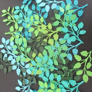 May include: Overhead view of paper cutouts of leafy branches in shades of green, teal, and olive green. The leaves are arranged in a circular pattern against a dark brown background. The leaves are various sizes and shapes.