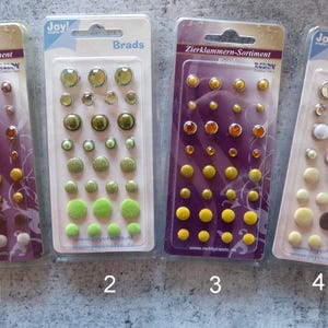 Könnte beinhalten: Vier Blisterpackungen mit sortierten Zierklammern, beschriftet mit Zierklammern-Sortiment und Brads. Jede Packung enthält mehrere Reihen kleiner, runder Metallklammern in verschiedenen Ausführungen, darunter metallische, matte und texturierte Oberflächen in Braun-, Grün- und Gelbtönen.