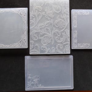 Könnte beinhalten: Eine Sammlung klarer Kunststoff-Prägeordner mit verschiedenen dekorativen Designs. Die Designs umfassen florale Muster, verzierte Bordüren und eine rechteckige Form mit einem Eckornament. Diese Ordner werden verwendet, um Papierarbeiten Textur zu verleihen.