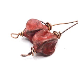 May include: Two irregular, glossy red ceramic beads secured with coiled copper wire. Each bead features a rounded, organic shape with deep indentations and a smooth, reflective surface. The copper wire wraps around the beads and extends outward.