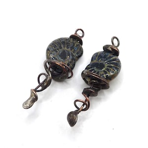 May include: Two pendants featuring dark, spiraled ammonite fossils encased in oxidized copper wire wrapping. Each fossil is topped with a small copper loop for hanging, and the wire extends downward into a decorative, coiled tail with a rounded tip.