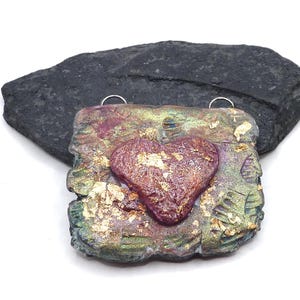May include: A square pendant featuring a raised, textured heart in a deep reddish-purple hue at the center. The surface is finished with a multi-colored metallic sheen of green, gold, and purple, accented with scattered gold leaf flakes throughout.