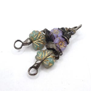 Pode incluir: Um par de brincos com argolas de metal em tom prateado escuro e detalhes em fio. Cada brinco inclui uma conta em forma de folha verde-clara com veios dourados e uma pequena conta em forma de flor roxa com um detalhe dourado no centro.
