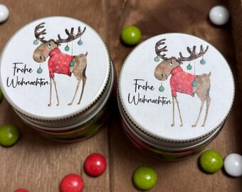 May include: Two glass jars with silver lids and white labels. The labels feature a cartoon moose wearing a red sweater and have the text "Frohe Weihnachten" on them.