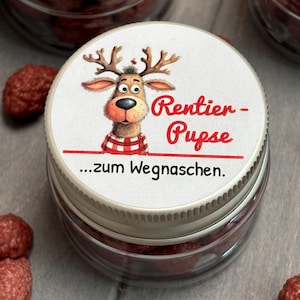 Könnte beinhalten: Klares Glas mit silbernem Deckel, gefüllt mit roten Süßigkeiten. Der Deckel zeigt ein Cartoon-Rentier mit dem Text "Rentier-Pupse" und "...zum Wegnaschen."