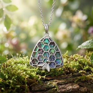 Puede incluir: Collar con colgante plateado con diseño de panal de abeja e incrustaciones de concha de abulón iridiscente. Un pequeño dije de abeja está unido a la parte inferior del colgante. El collar se exhibe sobre musgo verde.