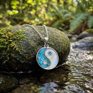 Könnte beinhalten: Eine silberne Halskette mit einem runden Anhänger, der das Yin-und-Yang-Symbol zeigt. Der Anhänger hat einen silbernen Rand, wobei eine Seite schillernde blau-grüne Abalone-Schale und die andere Seite weiße Perlmutt zeigt. Die Halskette wird auf einem moosbewachsenen Stein präsentiert.