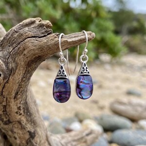 May include: A pair of drop earrings featuring teardrop-shaped iridescent purple and blue abalone shell inlays set in polished silver-toned metal. Each earring has a decorative triangular silver cap with an intricate cutout pattern and a hook.
