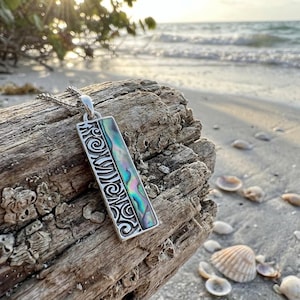 May include: A silver pendant necklace featuring a rectangular abalone shell inlay. The pendant has a silver frame with an intricate scroll design on one side. The necklace is displayed on driftwood on a beach.