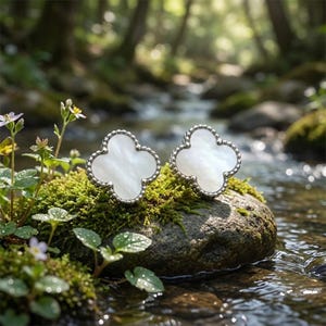 Peut inclure: Deux boucles d'oreilles argentées en forme de trèfle avec un centre en nacre blanche. Les boucles d'oreilles sont posées sur un rocher recouvert de mousse près d'un ruisseau, avec un arrière-plan flou de forêt. Bordure perlée.